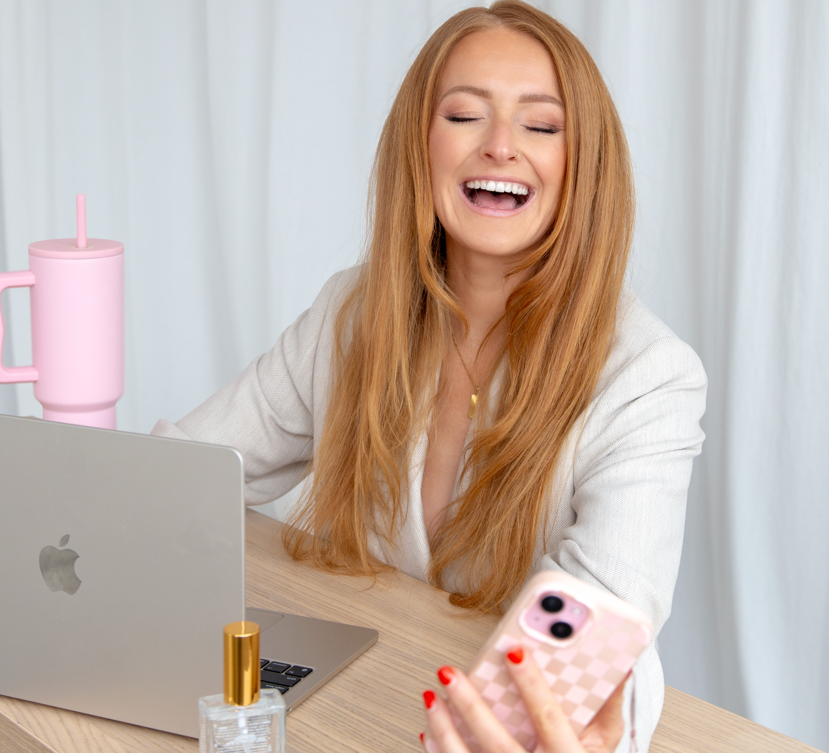 Woman sitting at a desk with a laptop, phone, and perfume bottle, laughing. apple computer. entrepreneur . red head. working