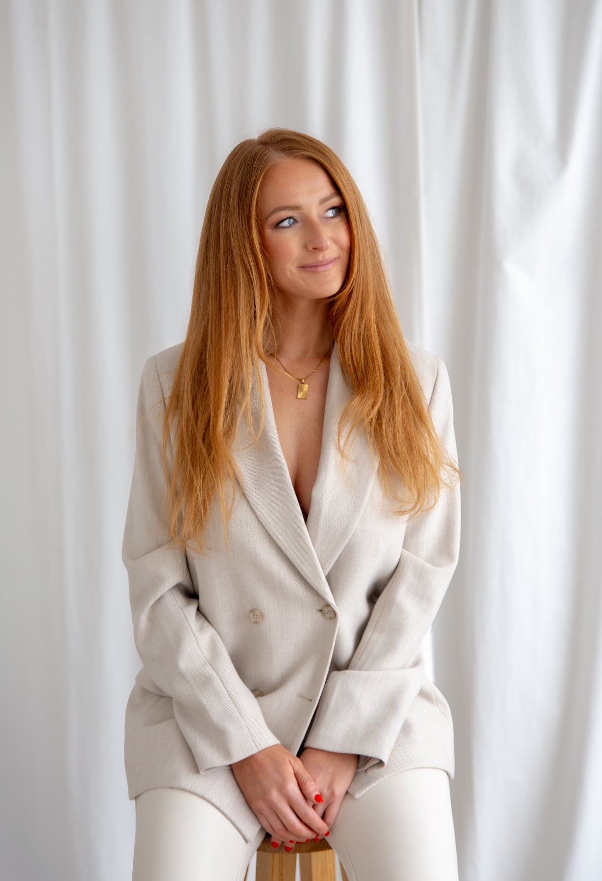 Woman in a light-colored suit sitting against a white curtain. Spray tan. women owned. small business. airbrush tan. salon. red head. entrepreneur
