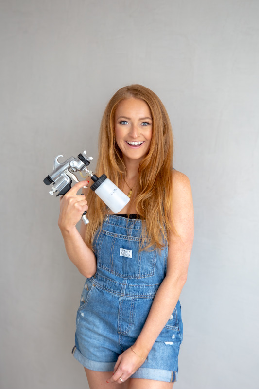 Woman holding a airbrush spray tan machine against a plain background.